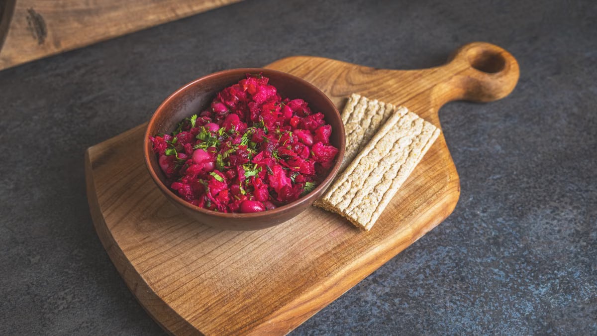 A vibrant beet salad served in a bowl with rye crackers on a wooden cutting board.